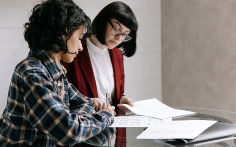 Dwie kobiety przeglądające i podpisujące dokumenty przy stole.