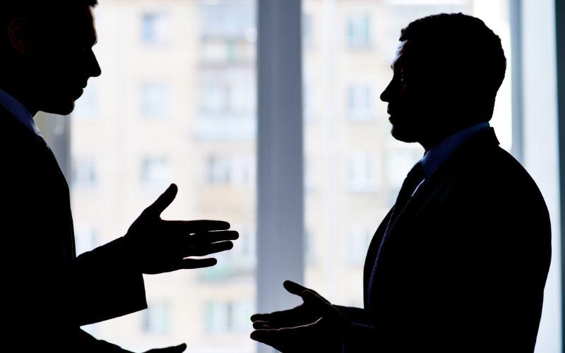 Sylwetki dwóch mężczyzn w garniturach rozmawiających na tle okna.