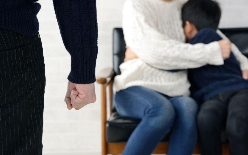 Mężczyzna z zaciśniętą pięścią, w tle matka przytulająca syna – dziecko jako świadek przemocy domowej