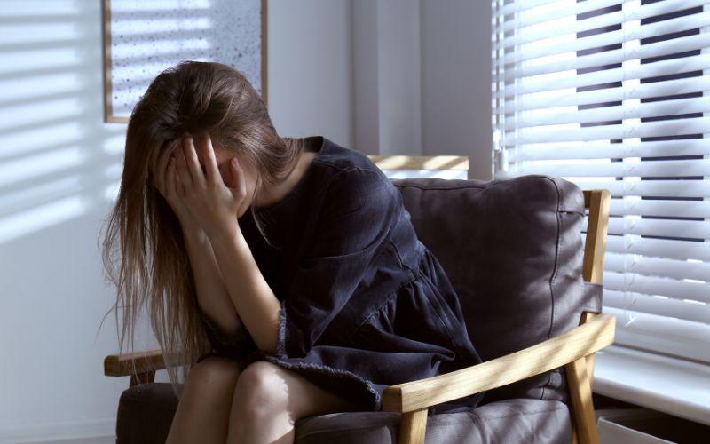 Kobieta siedząca samotnie w fotelu z twarzą ukrytą w dłoniach – skutki psychiczne znęcania się